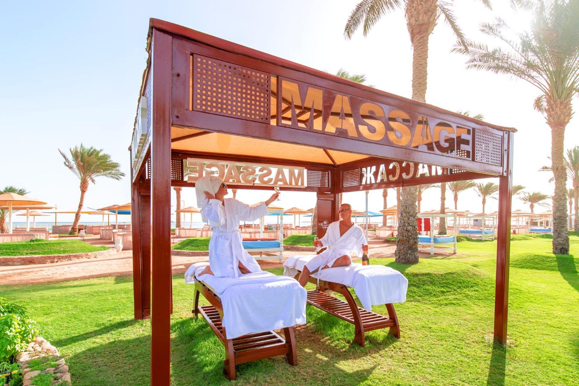منتجع رويال الباتروس مودرنا - للعائلات والأزواج فقط شرم الشيخ المظهر الخارجي الصورة The photo shows a massage area set up in a sunny outdoor environment. There are two massage tables under a wooden canopy with the word "MASSAGE" prominently displayed. Two individuals, dressed in white robes, appear to be engaged in a massage session