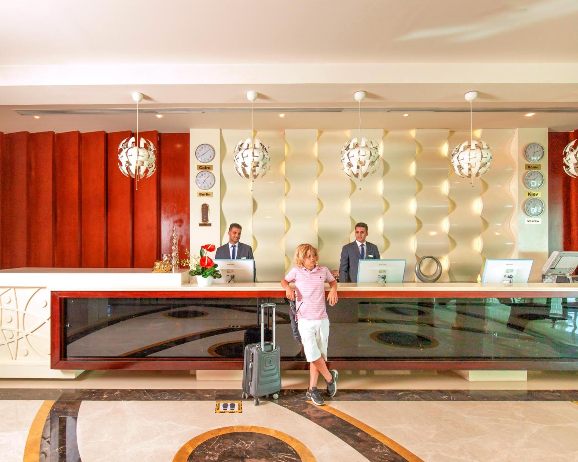 منتجع رويال الباتروس مودرنا - للعائلات والأزواج فقط شرم الشيخ المظهر الخارجي الصورة The photo depicts a hotel reception area. At the front desk, two staff members in suits are standing, presumably ready to assist guests. A man in casual attire, holding a suitcase, is standing in front of the desk. The reception has a modern design w