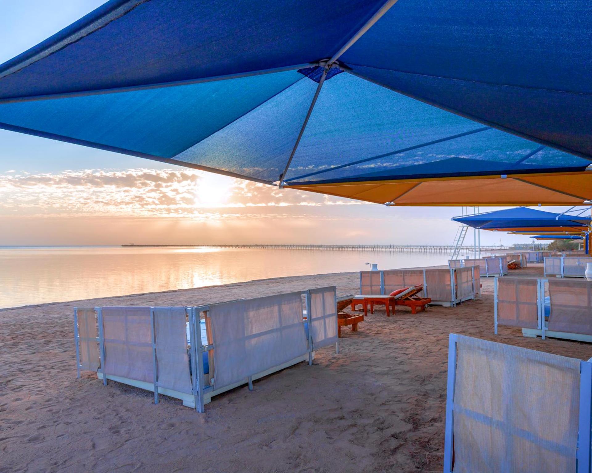 منتجع رويال الباتروس مودرنا - للعائلات والأزواج فقط شرم الشيخ المظهر الخارجي الصورة The photo shows a beach scene during sunset. There are colorful umbrellas in various shades of blue and orange set up over sandy areas. In the foreground, there are some wooden lounge chairs, and behind them, partition-like structures or beach cabana